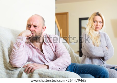 Portrait of a couple on a couch indoors after a quarrel. Sad husband sitting turned away from his wife that is looking at him in disappointment