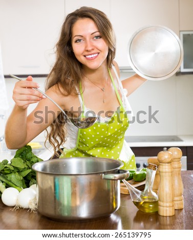 Smiling young housewife in apron cooking vegetable soup in home kitchen