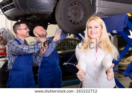 Happy woman at a car repair shop showing thumbs up while mechanics are examining her car\'s suspension in the background