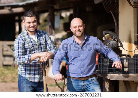 Two smiling male gardeners with farm tools near shed at sunny day