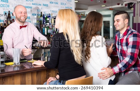 Portrait of group of people at the bar. A couple and alone attractive woman are sitting at the bar while the smiling bartender is pouring cocktail in a glass