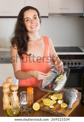 Smiling brunette woman putting pieces of lemon in fish at home kitchen