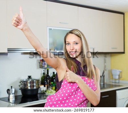 Cheerful young blonde woman turned up the thumbs in kitchen