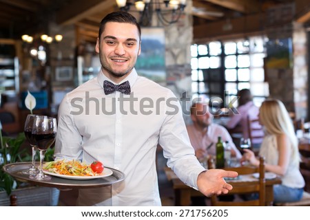 Happy smiling handsome waiter in a restaurant holding a tray with a salad and glasses full of wine in a restaurant