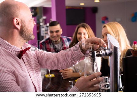 Bartender at a bar is pouring a beer from a tap and cheering up smiling visitors at a barstand