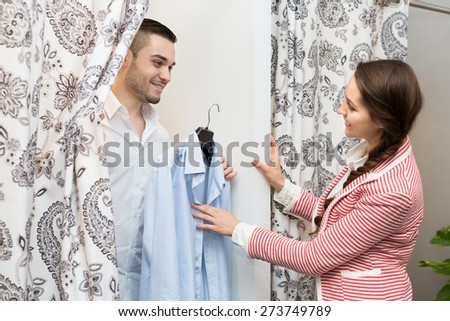 Portrait of smiling young couple with new apparel at fitting-room in clothing store. Focus on guy