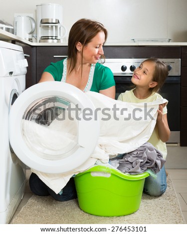 Home family laundry. Smiling mother with little daughter loading clothes into washing machine in kitchen. Focus on woman
