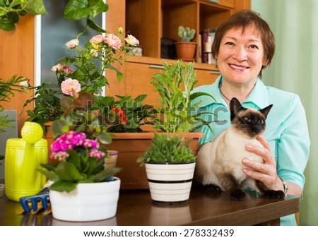 Smiling positive aged woman sitting at the table with cat and flower plants