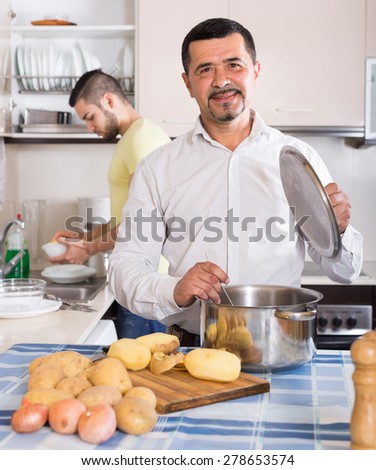 Two smiling male friends cooking with potato and doing dishes at home kitchen