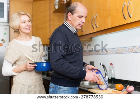 Elderly husband helping positive wife to cook in kitchen