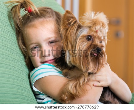 Happy blonde little girl embracing Yorkshire Terrier on sofa and smiling indoor