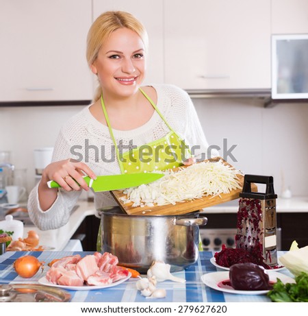 Cheerful young woman chopping cabbage for Russian soup at kitchen table