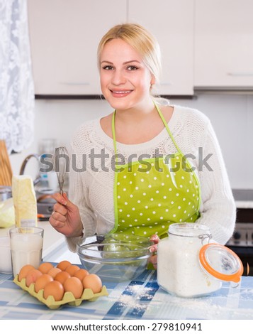 Happy smiling blonde housewife making dough with eggs and milk in kitchen