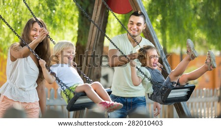 Happy young cheerful family of four at playground\'s swings. Focus on woman