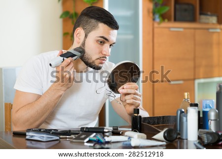 young american guy looking at mirror and shaving beard with trimmer