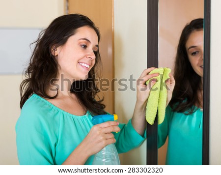Smiling brunette woman cleaning mirror with detergent at home