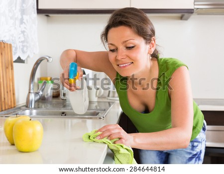 Cheerful young woman cleaning furniture with rag at home kitchen
