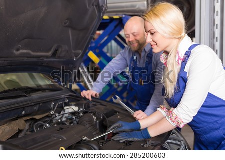 Auto service center crew with young smiling female repairing a broken car at garage. Focus on girl