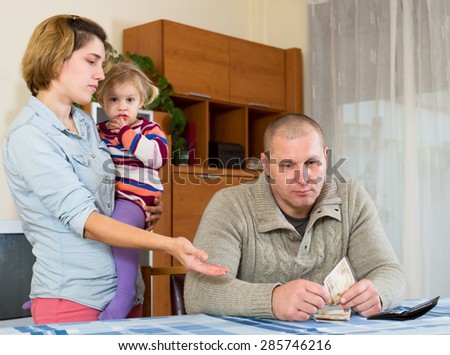 Wife nags his husband about money. She is holding their daughter on her arms he is sitting at the table with cash in his hands indoors