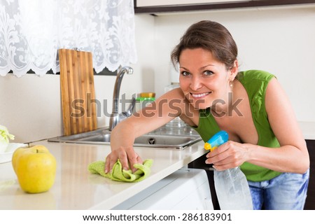 Cheerful smiling young housewife cleaning furniture with rag at home kitchen