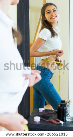 Young smiling girl measuring waist by centimeter in front if the mirror