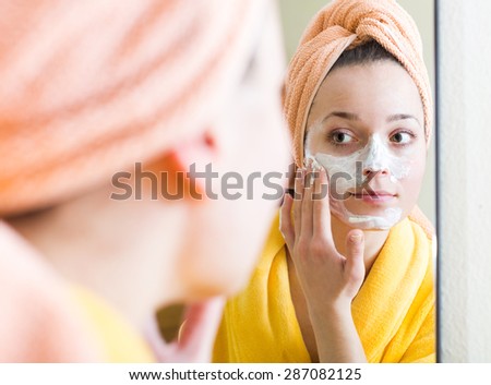 Young woman in bathrobe applying face pack indoors