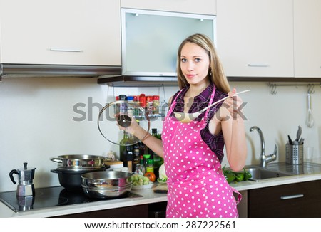 Smiling blonde young woman in apron with ladle testing soup from pan in domestic kitchen