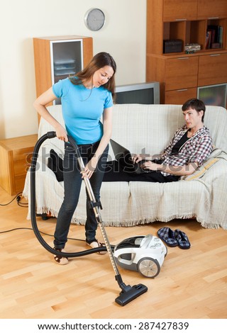 Girl cleaning with vaccuumcleaner at home while young man with notebook resting over sofa