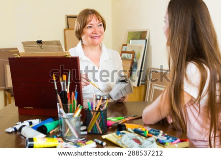 Mature female artist painting portrait of young girl at art studio with pencil and paints