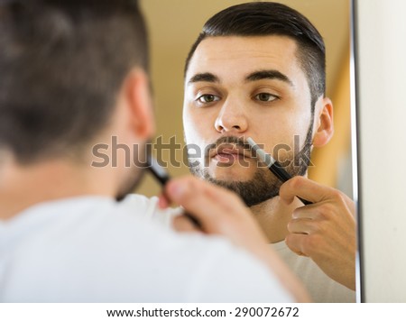 young american man looking at mirror and shaving beard with trimmer