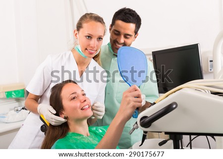 Dentist with assistant and contented female patient smiling to mirror at dental clinic