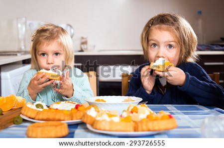 Two healthy young girls eating sweet cakes with cream. Their mouths are full and smeared with cream