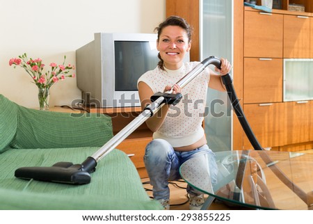 Smiling housewife cleaning sofa with vacuum cleaner at living room