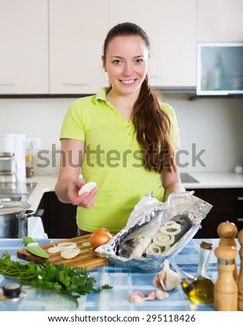 Smiling woman putting pieces of lemon in fish at home kitchen