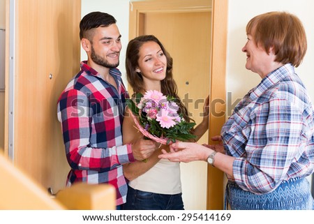 Senior woman meeting  smiling young couple with flowers at the door