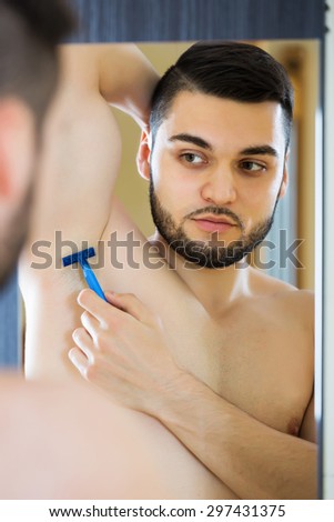 Young man looking at mirror and shaving armpit hair with razor