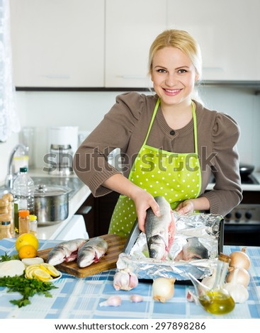 Happy blonde girl cooking fish  in frying pan at home kitchen