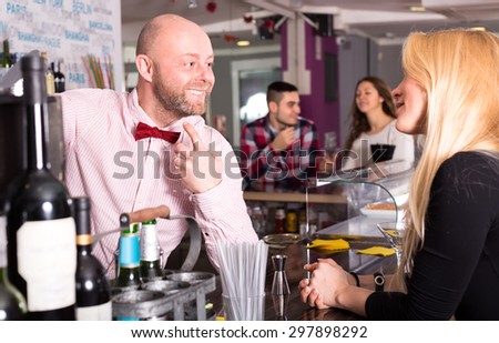 Smiling barman at the bar talking to a beautiful woman visitor another talking pair of visitors a man and a woman is visible in the distance