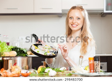 Smiling woman putting pieces onion to fish in frying pan at home kitchen