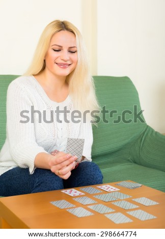 Happy young blonde sitting on a couch by the table and playing a solitaire card game