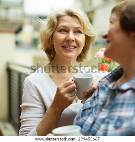 Smiling aged female friends staying on balcony with cup of coffee in hands