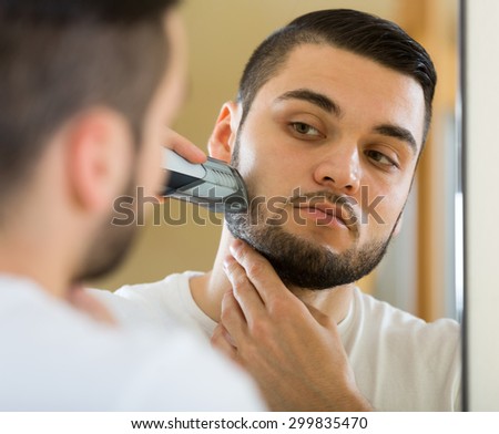 Young man looking at mirror and shaving by electric razor