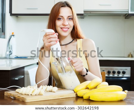 Positive woman making beverages with blender from bananas and milk at domestic kitchen