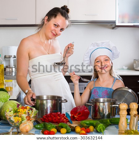 Smiling little girl and her mom cooking vegetable soup at home kitchen