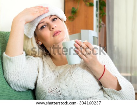 Feeling sick woman is sitting on a sofa at home with a wet cold towel on her forehead and a cup of warm drink in her hand