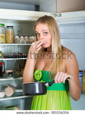 Blonde young woman holding her nose because of bad smell near fridge at home