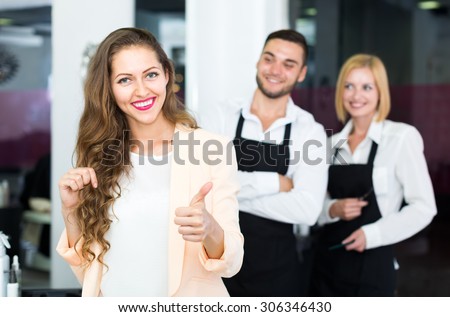 Happy woman standing in a hair salon touching her hair and showing thumbs up sign while hairdressers are standing in the back