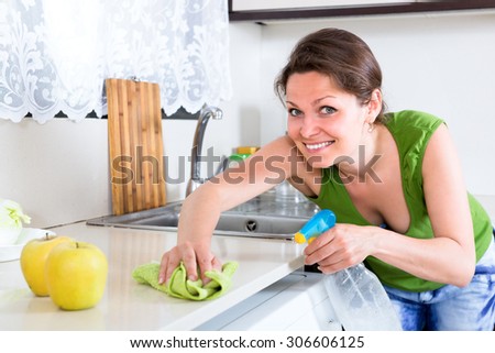 Woman with a rag and clear bottle with cleaning substance is cleaning kitchen furniture from dust and dirt