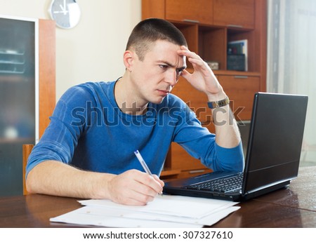 Sad frustrated guy looking at financial documents in laptop in home interioÃ�Âº sitting by table