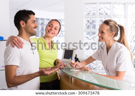 Positive young couple talking with smiling secretary at reception in the clinic